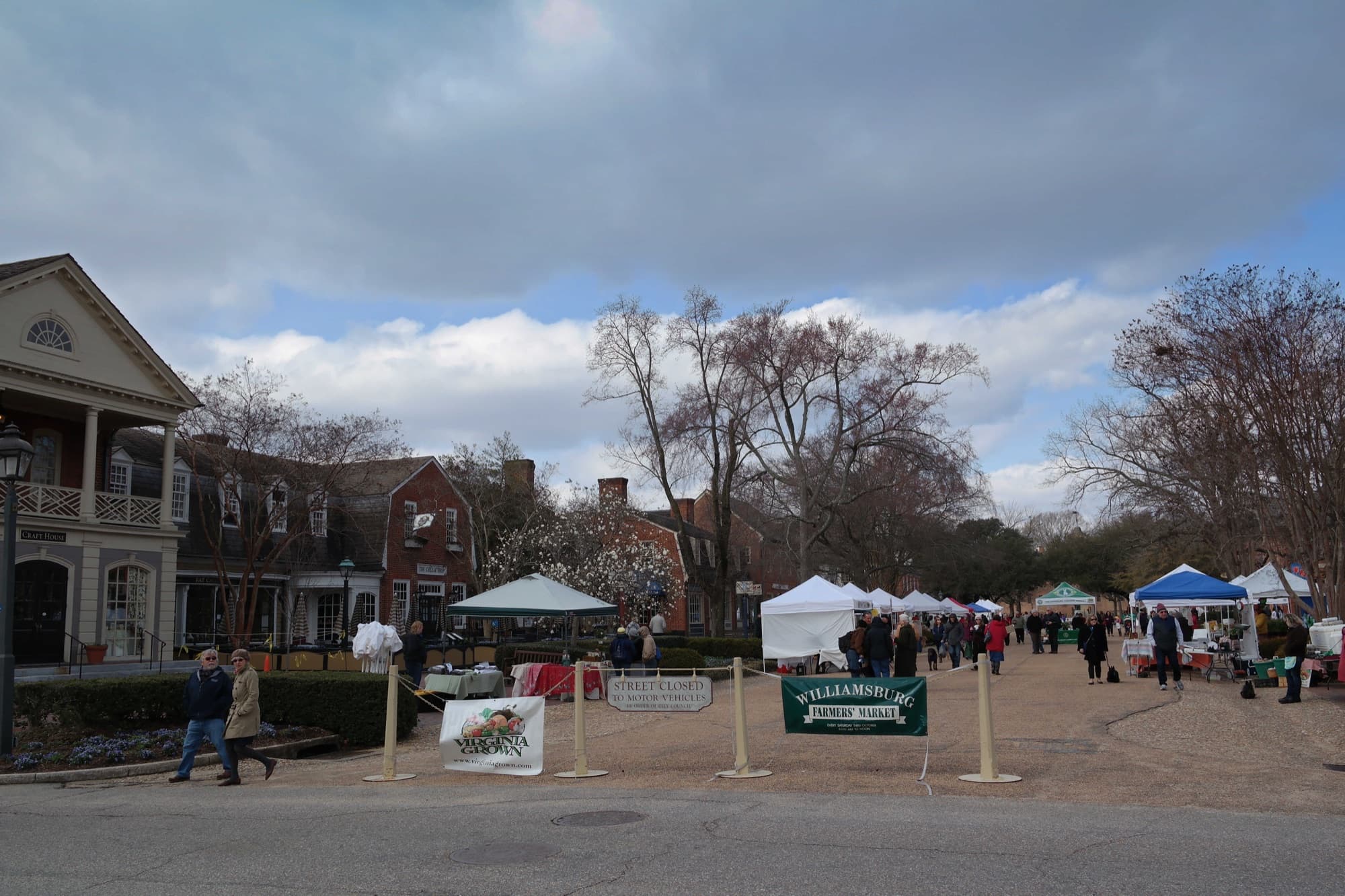 Shoppers browsing produce stalls under trees at a farmers market in Merchants Square, Williamsburg, Virginia.
